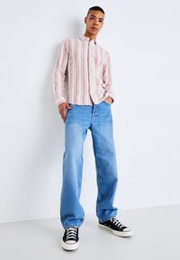 Chemise à rayures, boutonnée, en tissu rose et blanc, associée à un jean large bleu clair et des baskets noires avec des semelles blanches.