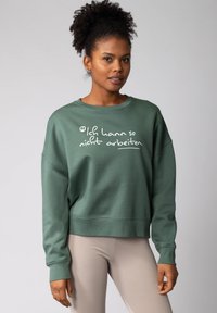 Sweatshirt vert avec texte blanc, poignets et ourlet côtelés, coupe décontractée. Stylisé avec des leggings côtelés légers, le modèle a des cheveux bouclés naturels.