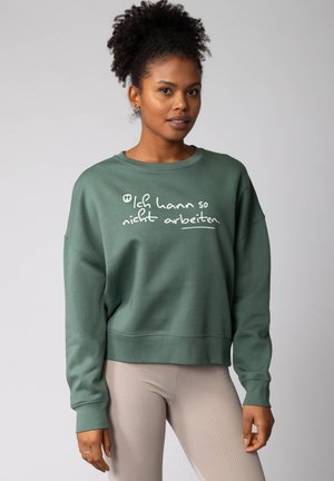 Groene sweatshirt met witte tekst, geribde manchetten en onderrand, relaxte pasvorm. Gestyled met licht geribde leggings, het model heeft natuurlijk krullend haar.