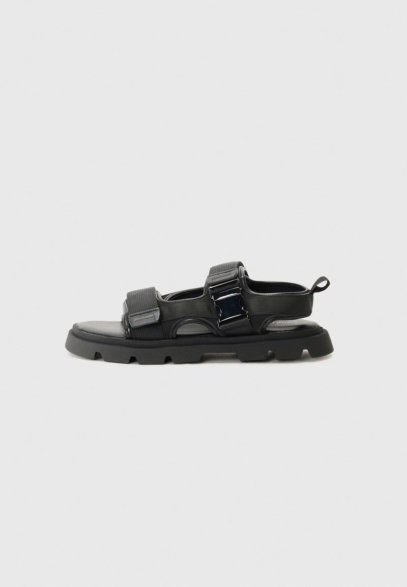 Black sandals with a textured fabric upper, wide straps, metal buckle accents, and a thick rubber sole featuring a lug pattern.