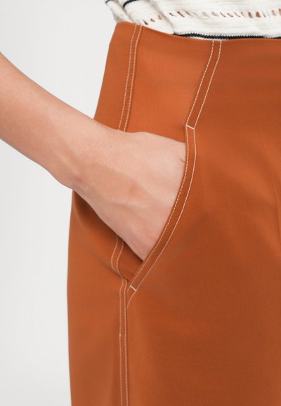 Rost-orange Hosen mit einer Seitentasche, die kontrastierender weißer Nähte aufweisen. Glatte Stofftextur mit tailliertem Schnitt und klaren Linien.