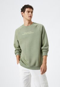 Sudadera verde hecha de un tejido suave con mangas raglán, puños acanalados y un escote en cuello redondo. Presenta el texto: "Las personas geniales son el mejor tipo de peligro."
