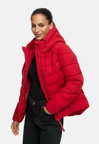 Punane puhv jacket, millel on kapuuts, tikandiga muster ja eesmine lukK. Kombineeritud musta topi ja tumedate teksadega, lõdva lõikega.