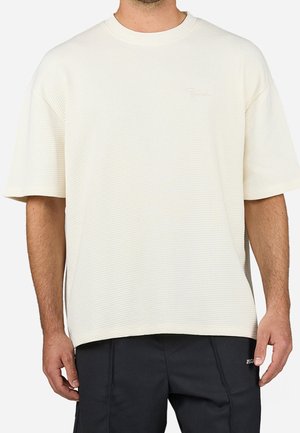 Mężczyzna w luźnej, kremowej, fakturowanej koszuli z krótkim rękawem i czarnych spodniach, stojący na białym tle.