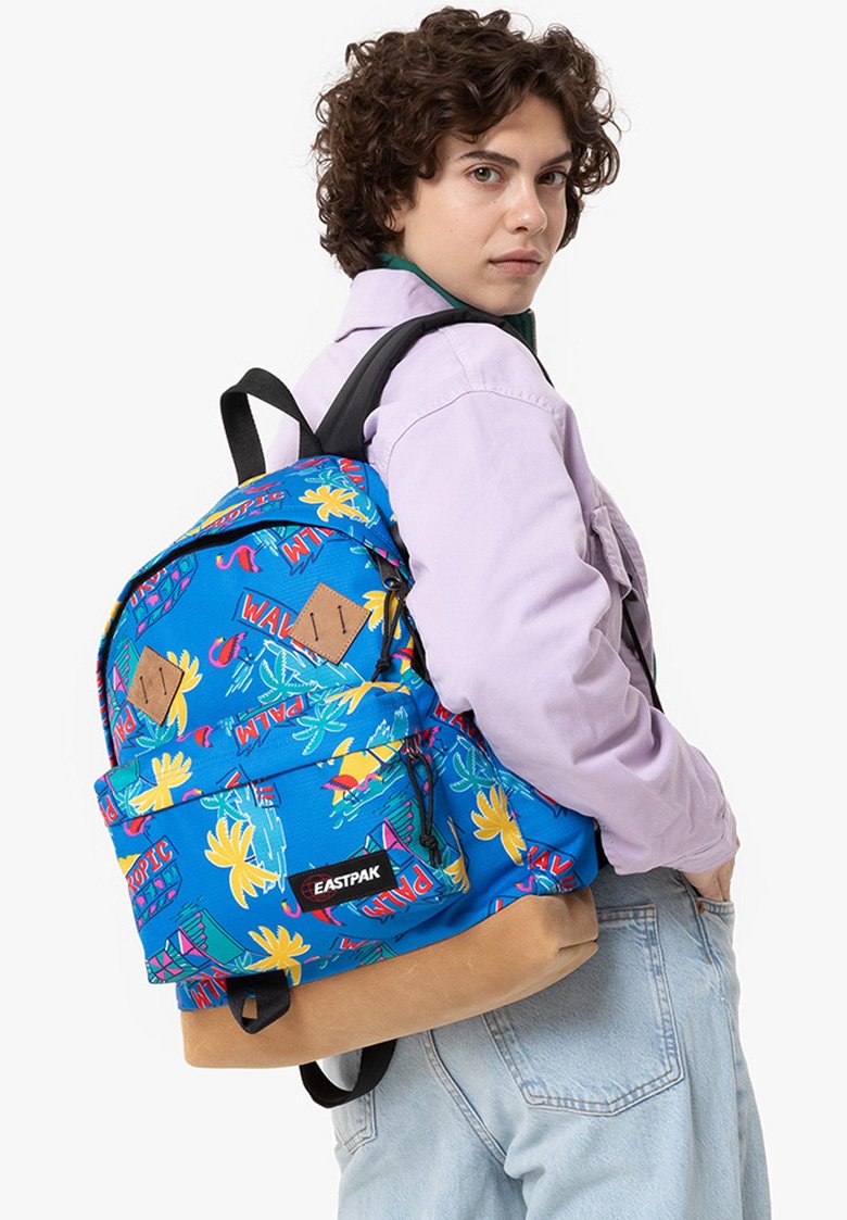 Persona con chaqueta morada clara y jeans azul claros lleva una mochila Eastpak de patrón tropical brillante en color azul sobre un hombro.
