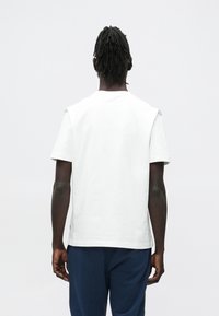 Moški s temno poltjo in dreadlocki, oblečen v preprosto belo majico s kratkimi rokavi in temno modre hlače, stoji z obrazom obrnjeno stran proti belemu ozadju.