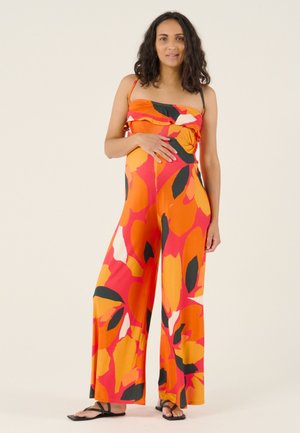  zwangere vrouw met schouderlang krullend haar, gekleed in een oranje, rode, zwarte en witte strapless jumpsuit met bloemenprint, staand met één hand op haar buik.