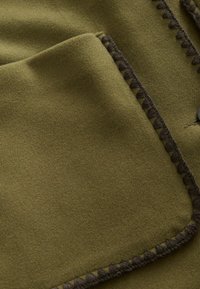 Olivroheline mantlikangas pehme, tekstuuriga pinnaga; tumeda pruuni tikandiga detailid servadel ja ühekordne nööp sulgemiseks.