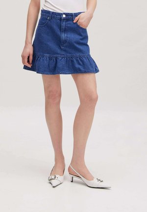 Frau trägt einen blauen Jeansrock mit Rüschensaum, weißes Top und weiße Slingback-Kitten-Heel-Schuhe, steht auf einem schlichten hellen Hintergrund.