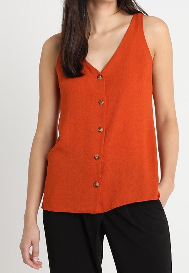 Femme portant un haut orange sans manches boutonné avec un col en V, associé à un pantalon noir, debout devant un fond clair uni.