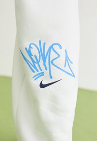 Hvidt stof med blåt graffiti-lignende tekst og et sort Nike swoosh logo trykt på det, mod en lys grøn og hvid baggrund.