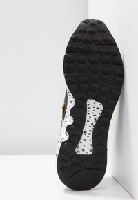 Semelle noire avec motifs antidérapants texturés, semelle intermédiaire blanche présentant un design tacheté, et une partie supérieure lisse noire et dorée.