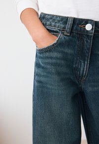 Tmavomodré rifle z denimu s voľným strihom, vybavené predným vreckom a kontrastným oranžovým prešitím. Pás má zapínanie na gombík.