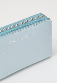 Gaiši zila ādas maciņš ar teksturētu virsmu, ar sudraba rāvējslēdzēju un iepresētu "Calvin Klein" logo. Taisnstūra forma.
