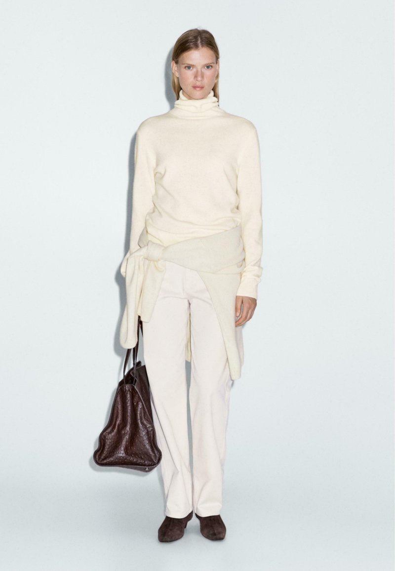 Maglione a collo alto crema, pantaloni chiari e borsa marrone. Tessuto morbido, vestibilità rilassata, con un dettaglio annodato sul maglione.