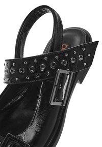 Sandale en cuir verni noir avec bout pointu, bride réglable, boucles métalliques et détails décoratifs à œillets le long de la bride.