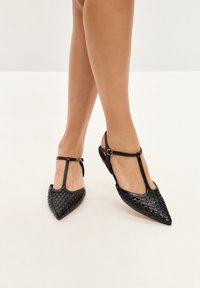 Pantofi din piele împletită neagră, cu vârf ascuțit și design T-strap. Include cataramă ajustabilă la gleznă și talpă plată.