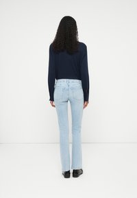 Une personne avec de longs cheveux bouclés se tient face à un mur blanc, portant un haut à manches longues bleu marine, un jean bleu clair et des chaussures noires.