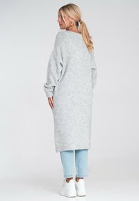 Kobieta z blond koński ogon, ubrana w długi szary sweter, jasnoniebieskie dżinsy, białe sneakersy i złote kolczyki w kształcie kół, stojąca przodem w lewo.
