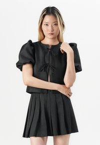 Svart cropped topp med puffermer og sløyfefestning, kombinert med et plisserte svart skjørt. Glatt stoff med en strukturert silhuett.