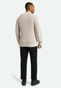 Pulover bej tricotat, cu manșete și tiv din tricot, combinat cu pantaloni negri și pantofi închiși la culoare; prezintă un guler înalt și mâneci lungi.