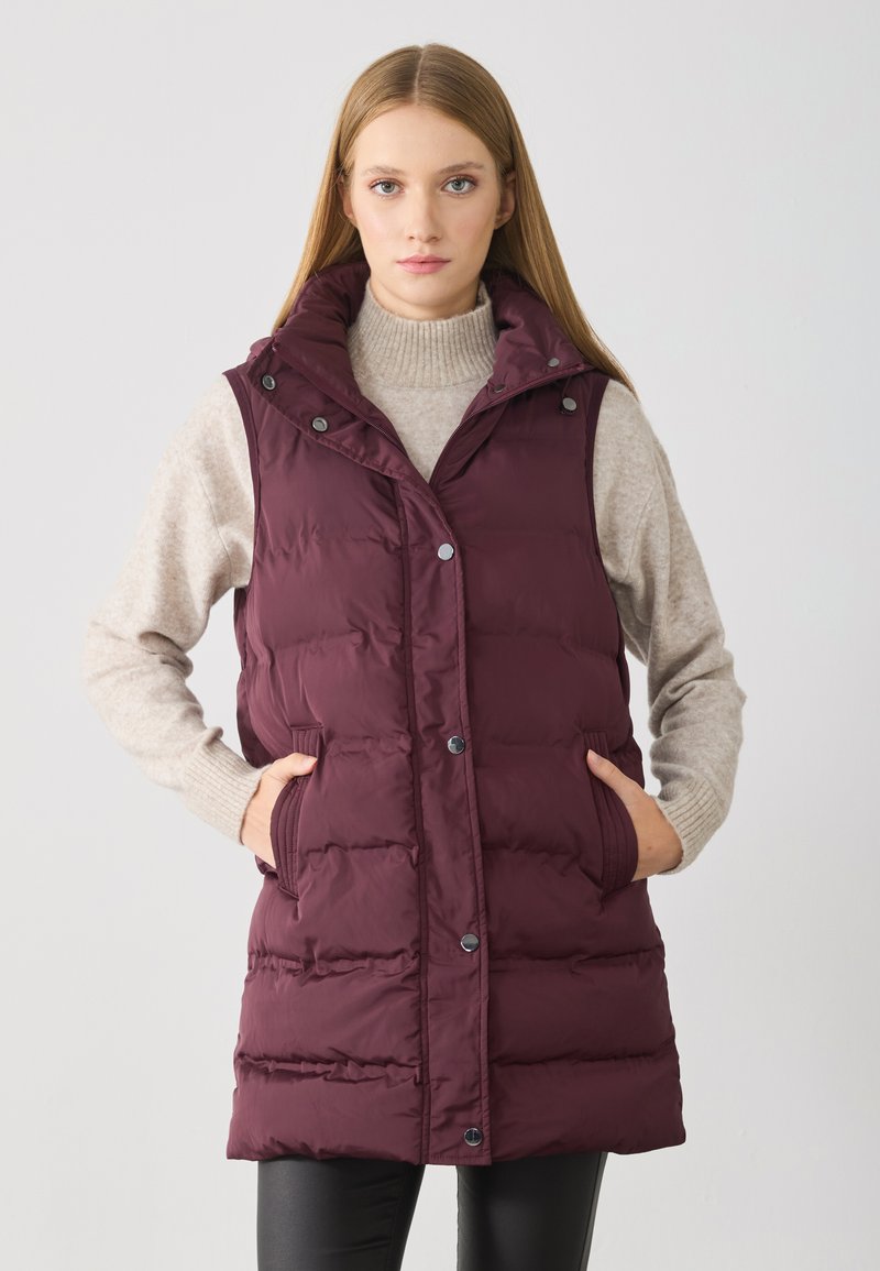 Bordeauxfarbener Puffer-Weste mit hohem Kragen, Druckknöpfen, seitlichen Taschen und gestepptem Design, getragen über einem hellbeigen Pullover.