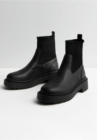 Stivali alla caviglia neri con tomaia in pelle testurizzata e parte superiore in maglia a costine simile a un calzino, suola spessa e linguette di trazione su uno sfondo bianco semplice.