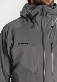 Homme portant une veste imperméable grise à capuche avec fermeture éclair frontale et poche poitrine, logo "Mammut" sur le côté gauche de la poitrine.