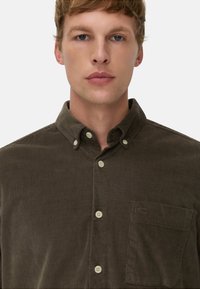 Koszula zapinana na guziki w ciemnooliwkowym korduroju, z nasadzaną kołnierzykiem, przednią kieszenią i beżowymi guzikami. Miękka tekstura i klasyczne elementy wzornictwa.