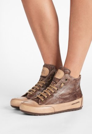 Sneakers alte in pelle marrone con punta, lacci e suola beige indossate a piedi nudi su sfondo bianco.