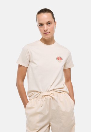 Femme avec un t-shirt beige clair arborant un petit motif coloré de panier de fruits sur la poitrine, assorti à un pantalon à cordon assorti, mains dans les poches.