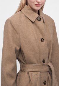 Femme portant un manteau en laine beige ceinturé avec un large col et des boutons marron foncé, debout devant un fond clair uni.