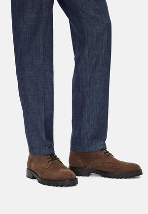 Rjavi semišasti čevlji z teksturirano površino, s vezalkami in robustnim črnim podplatom, kombinirani s temno modrimi hlačami.