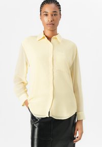 Camicia giallo chiaro, semitrasparente, a maniche lunghe con chiusura a bottoni e taschino sul petto, abbinata a una gonna di pelle nera.