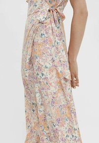 Robe portefeuille à motifs floraux dans des tons pastel d'orange, de violet et de bleu, nouée avec un nœud à la taille, présentée sur le côté d'une personne debout.