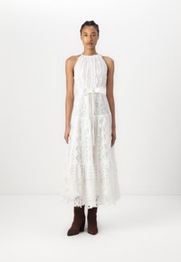 Une robe en dentelle blanche avec un col halter et un n�œud en ruban à la taille, dotée d'une jupe évasée et de motifs complexes. Le mannequin porte des bottes marron.
