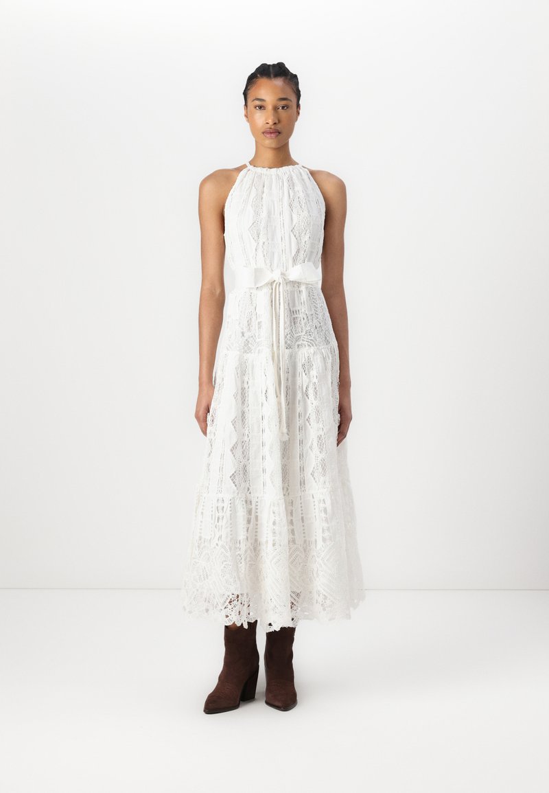 Une robe en dentelle blanche avec un col halter et un nœud en ruban à la taille, dotée d'une jupe évasée et de motifs complexes. Le mannequin porte des bottes marron.