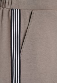 Beige sportiniai kelniukai su tekstūruotu paviršiumi, turintys juodą šoną su trimis baltomis linijomis ir šoniniu kišeniu su paslėptu atidarymu.