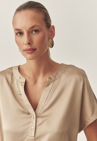 Beige satinblus med V-ringning och korta ärmar, med subtila knapppdetaljer och en slät, glansig yta. Guldiga statementörhängen.