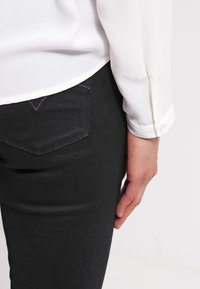 Personne portant une chemise blanche à manches longues et un pantalon noir, montrant le côté droit de la taille à la main sur un fond uni.