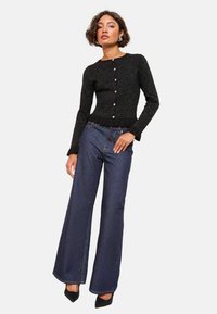 Černý texturovaný cardigan s peplum pasem a knoflíkovým zapínáním, sladěný s tmavomodrými džíny s vysokým pasem a černými podpatky.
