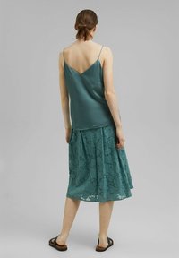 Top de satén verde azulado con un escote en V en la parte posterior, combinado con una falda de encaje de largo hasta la rodilla con patrones florales. Sandalias completan el conjunto.