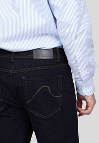 Jeans en denim foncé avec des coutures jaunes, dotés d'une étiquette en cuir à l'arrière. Chemise de dress bleu clair et blanche à rayures visible par-dessus les jeans.