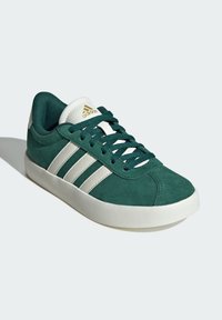 Zapatilla de ante verde con diseño de tres rayas blancas, suela blanca, acentos de logo dorado y cordones texturizados. Punta redondeada.