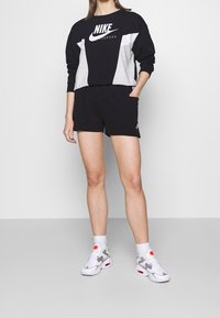 Person klädd i svart, grått och vitt Nike-tröja och svarta shorts med vita strumpor och vita Nike-sneakers, stående framför en enfärgad bakgrund.