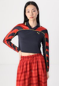 Svart og rødt langermet crop top med bølgete mønster, gule detaljer og polo-krage, sammen med et rødt skjørt med rutete mønster.