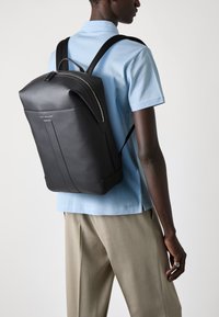 Black leather backpack with a sleek, structured design, featuring a curved zipper and adjustable straps. Worn by a figure in a light blue shirt.