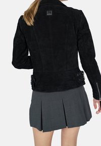 Schwarze Wildlederjacke mit tailliertem Design, ausgestattet mit einem Logo-Patch, seitlichen Schnallen und strukturiertem Oberflächen. Kombiniert mit einem grauen Faltenrock.
