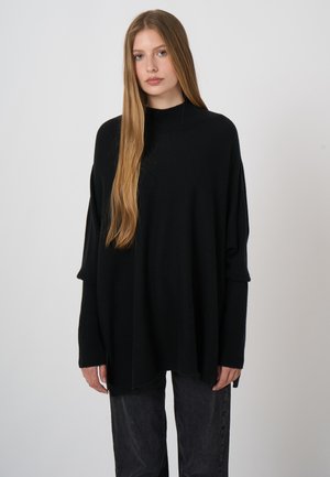Junge Frau mit langen blonden Haaren, die einen übergroßen schwarzen Pullover und dunkelgraue Hosen trägt, stehend vor einem schlichten hellen Hintergrund.
