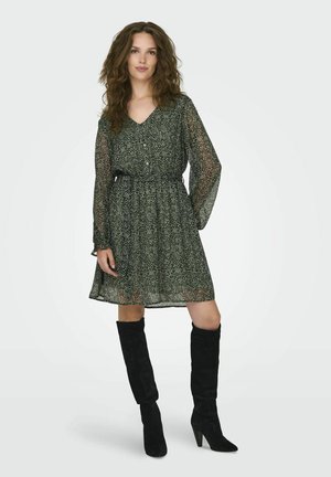 Robe fluide verte avec un décolleté en V, des manches longues, une taille froncée et un motif subtil. Associée à des bottes en daim noires montantes.